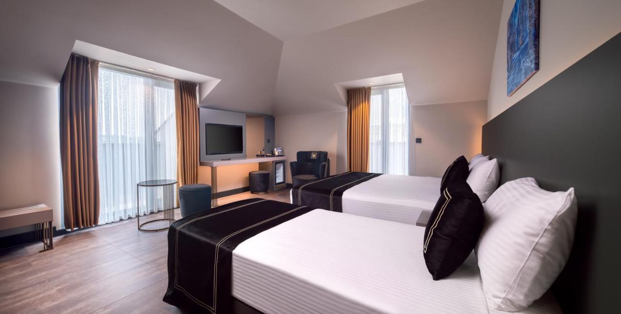 Menalo Hotel Premium Istanbul Airport Exterior photo The photo shows a modern hotel room featuring two beds with white linens and black throw pillows. There is a television mounted on the wall opposite the beds. A small seating area includes a comfortable chair and a table, while a mini-fridge and a co