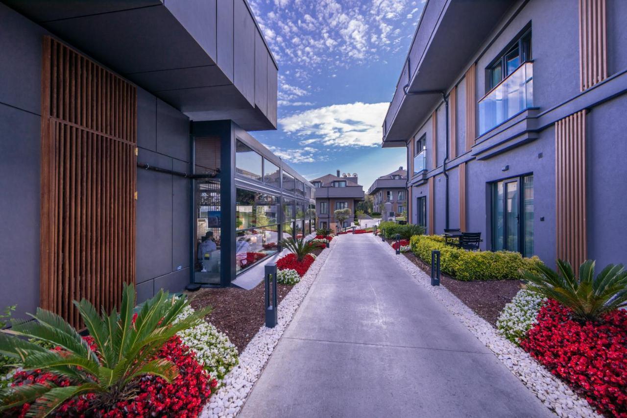 Menalo Hotel Premium Istanbul Airport Exterior photo The photo depicts a well-designed exterior pathway leading between modern buildings. The pathway is bordered by lush greenery and vibrant flower beds featuring a mix of red, white, and green plants. The sky above is partly cloudy, adding a serene bac