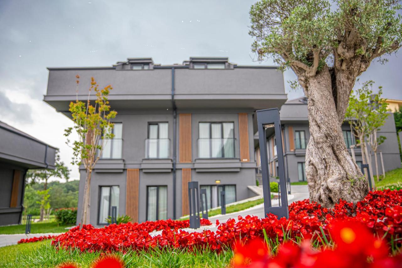 Menalo Hotel Premium Istanbul Airport Exterior photo The photo features a modern, two-story building set within a landscaped area. The building has a sleek design with large windows and a neutral color palette, primarily in shades of gray and brown. In the foreground, there's a vibrant display of red f