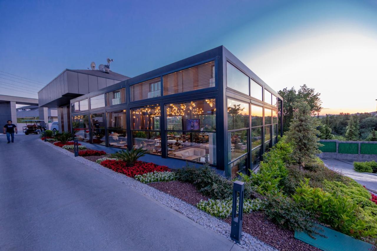 Menalo Hotel Premium Istanbul Airport Exterior photo The photo showcases a modern building with large glass windows, highlighting its sleek architectural design. The structure appears to be situated in a landscaped area, featuring vibrant flower beds and greenery. The setting is during the evening, as 