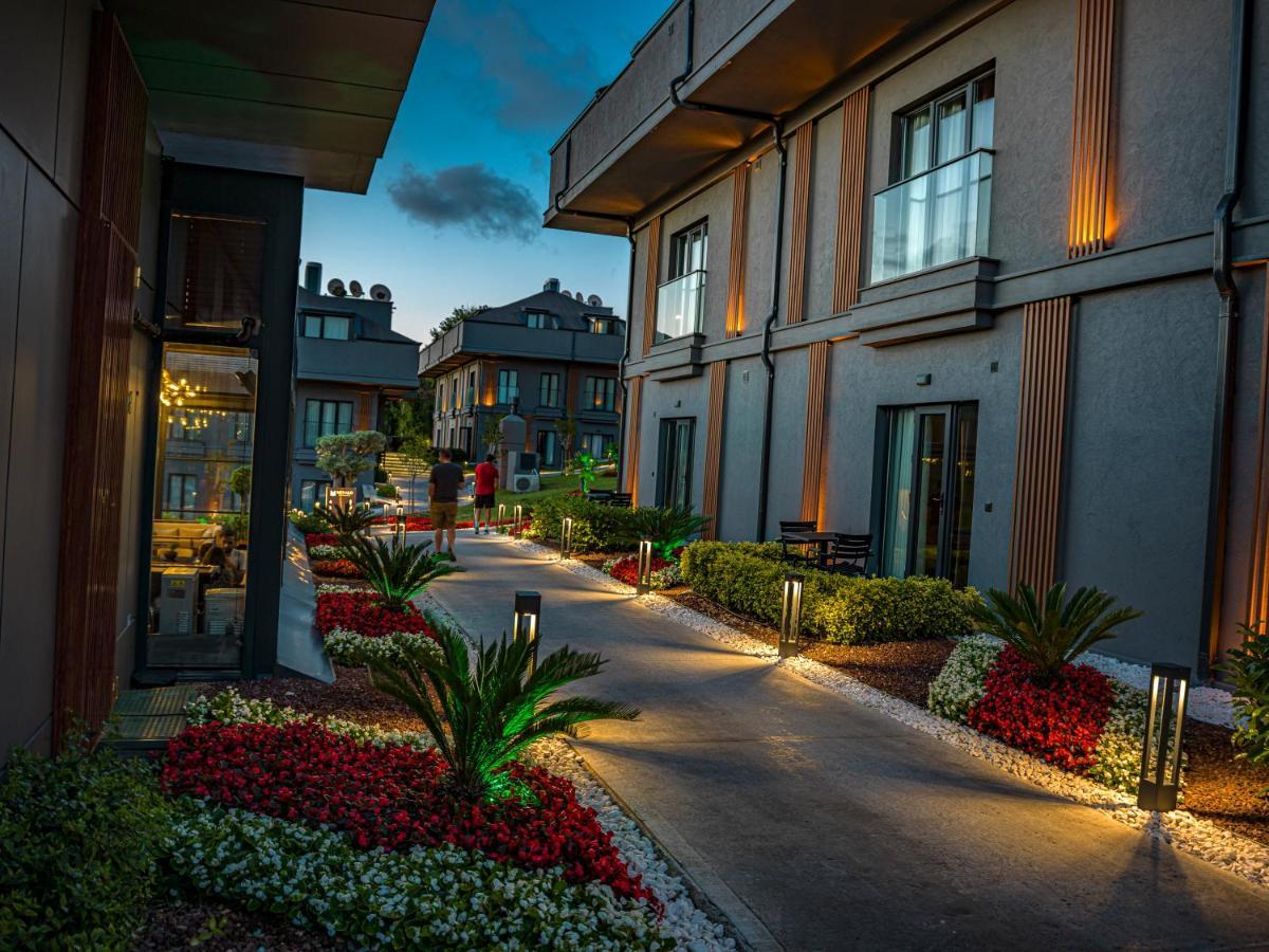 Menalo Hotel Premium Istanbul Airport Exterior photo The photo shows a pathway between modern buildings, illuminated by soft lighting. The path is lined with colorful flower beds featuring red and white blooms and small palm plants, adding to the aesthetic appeal. The buildings are designed with a mix 