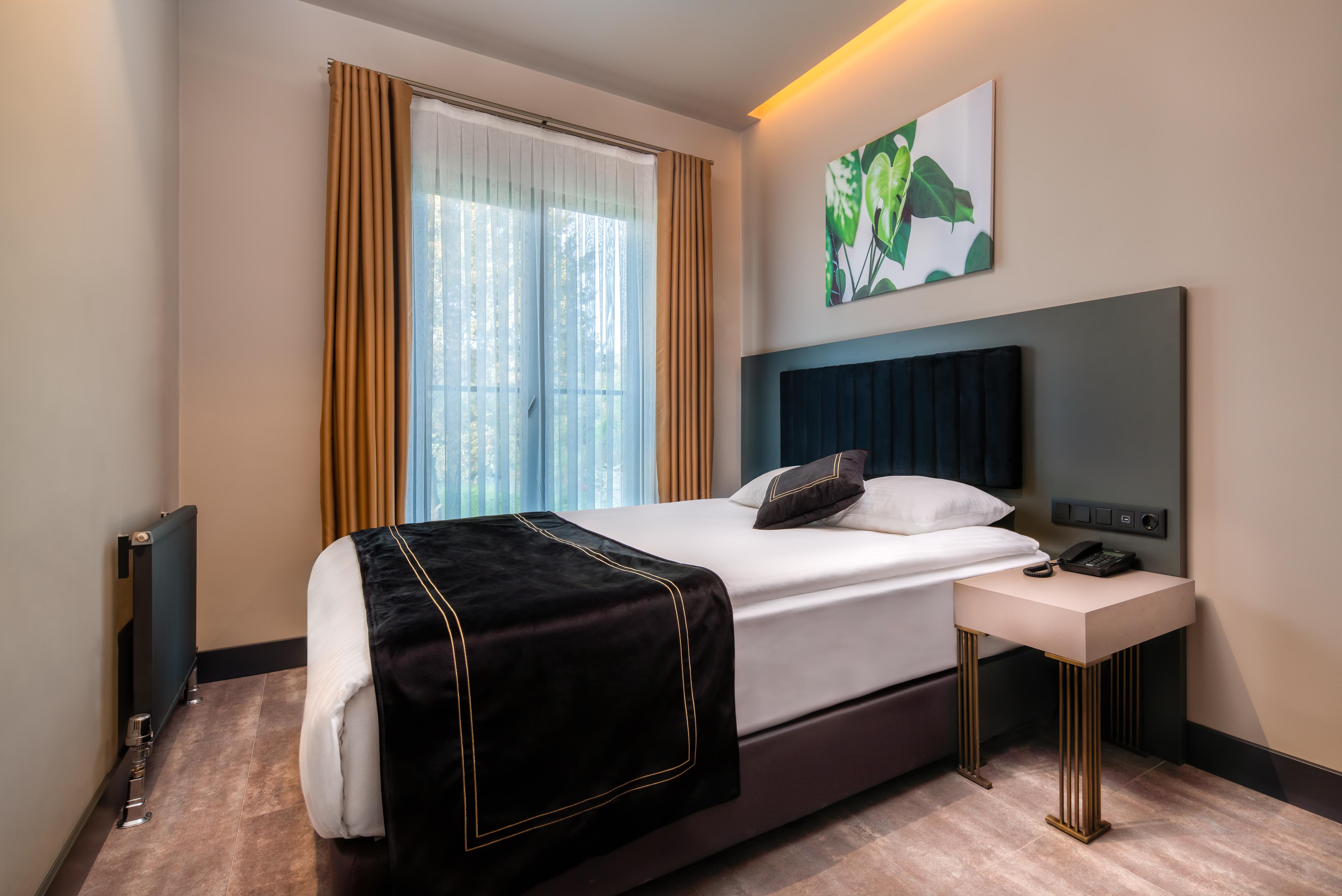 Menalo Hotel Premium Istanbul Airport Exterior photo The photo shows a modern hotel room featuring a neatly made bed with a black and white bedspread. The headboard is upholstered in a dark fabric. There's a small bedside table beside the bed with a lamp or phone. The room has large windows covered wit