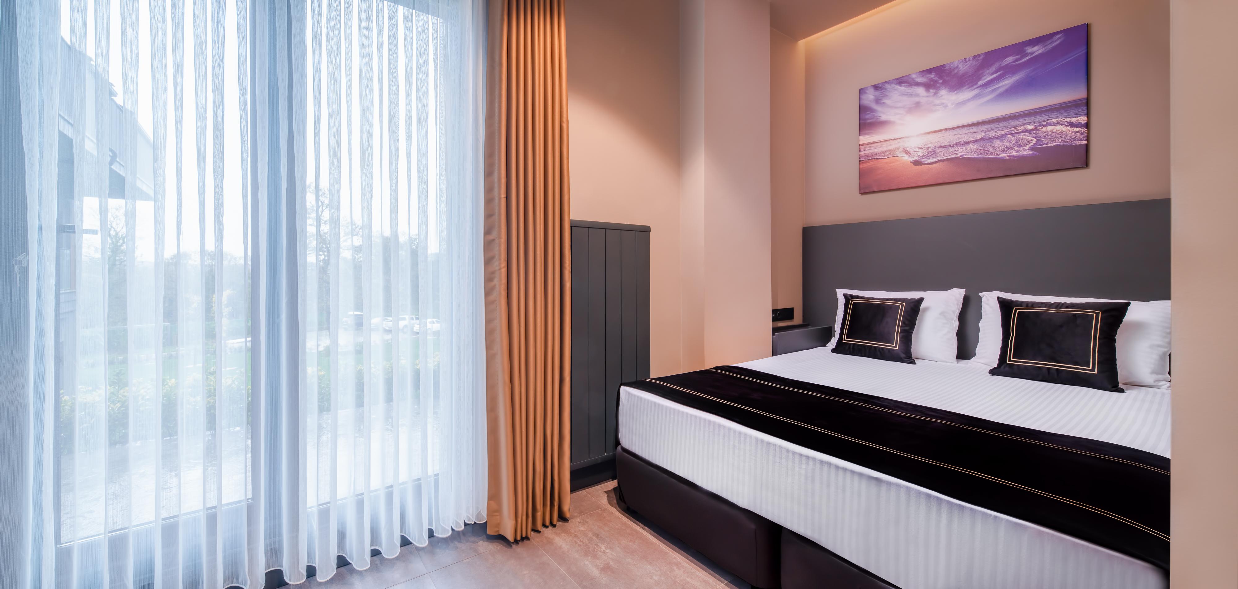 Menalo Hotel Premium Istanbul Airport Exterior photo The photo displays a modern hotel room featuring a double bed with white linens and dark accents. There are two decorative pillows on the bed. To the left, large windows draped with sheer white curtains allow natural light to filter in, complemented 