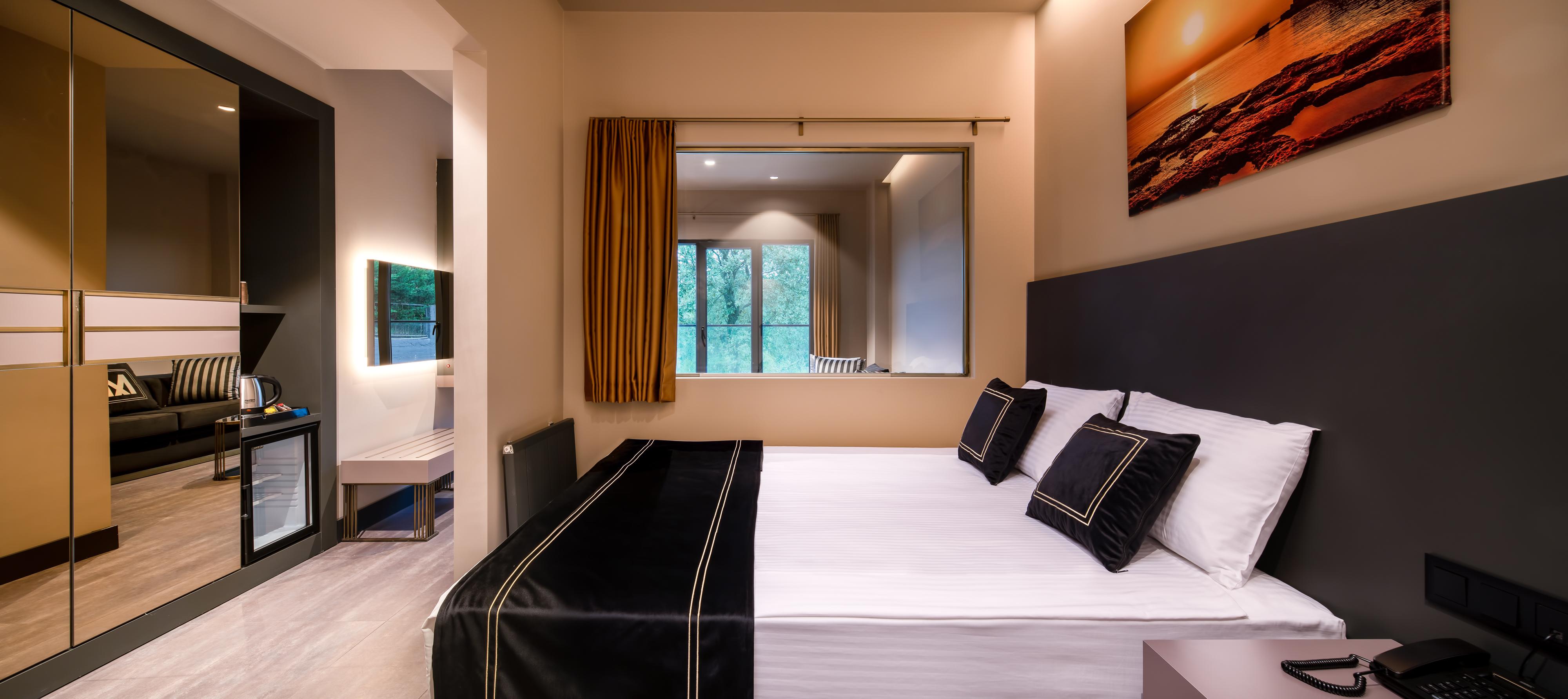 Menalo Hotel Premium Istanbul Airport Exterior photo The photo shows a cozy hotel room featuring a double bed with white linens and dark-colored decorative pillows. There is a large window with bright yellow curtains that lets in natural light, revealing a view of greenery outside. On the wall opposite