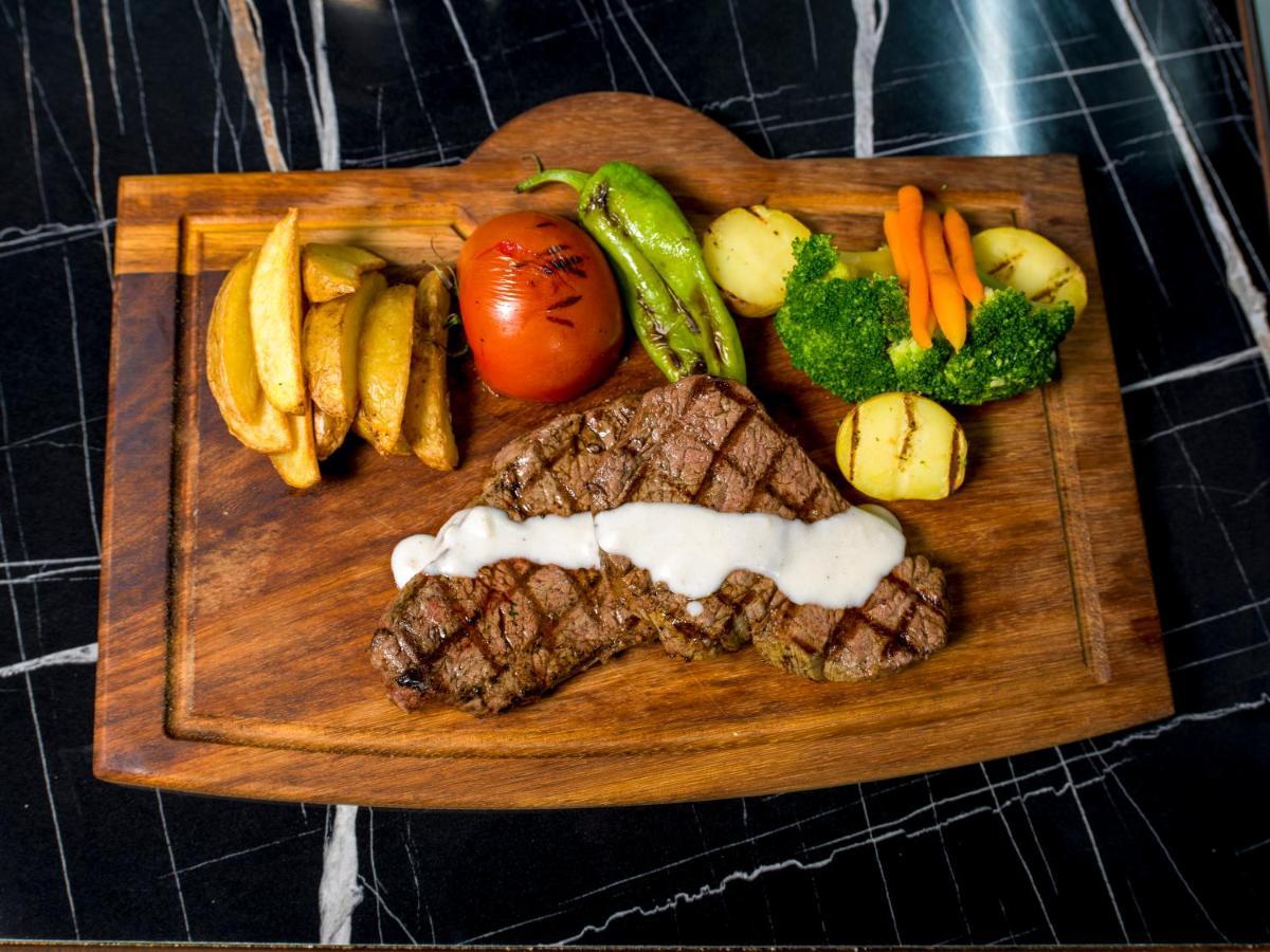 Menalo Hotel Premium Istanbul Airport Exterior photo The photo shows a meal presented on a wooden cutting board. The main feature is a grilled steak, which has grill marks and is topped with a creamy sauce. Accompanying the steak are several side items: 

- A portion of golden-brown potato wedges
- A w