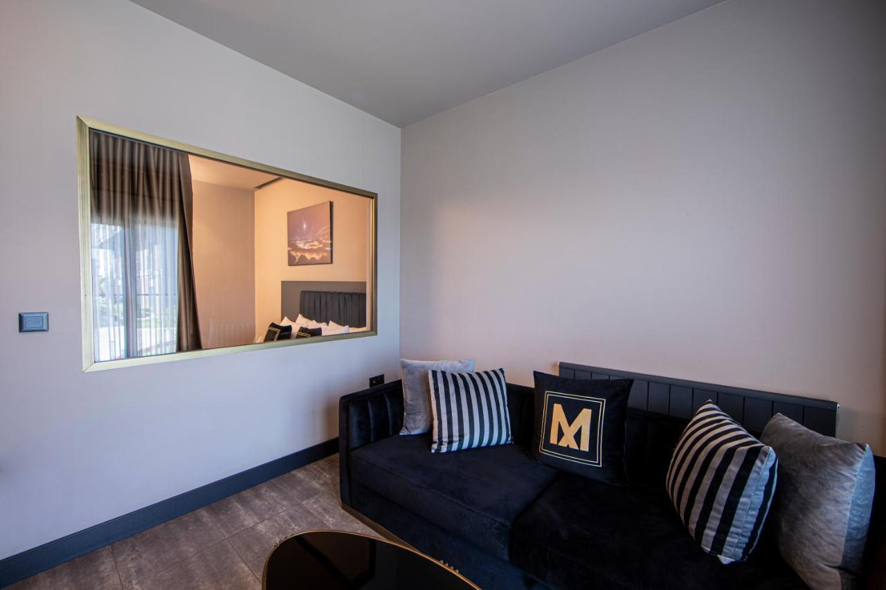 Menalo Hotel Premium Istanbul Airport Exterior photo The photo shows a modern living space featuring a stylish black sofa adorned with striped cushions, one of which has an "M" design. There is a large mirror framed in gold on the wall, reflecting part of the room. The wall opposite the sofa appears to