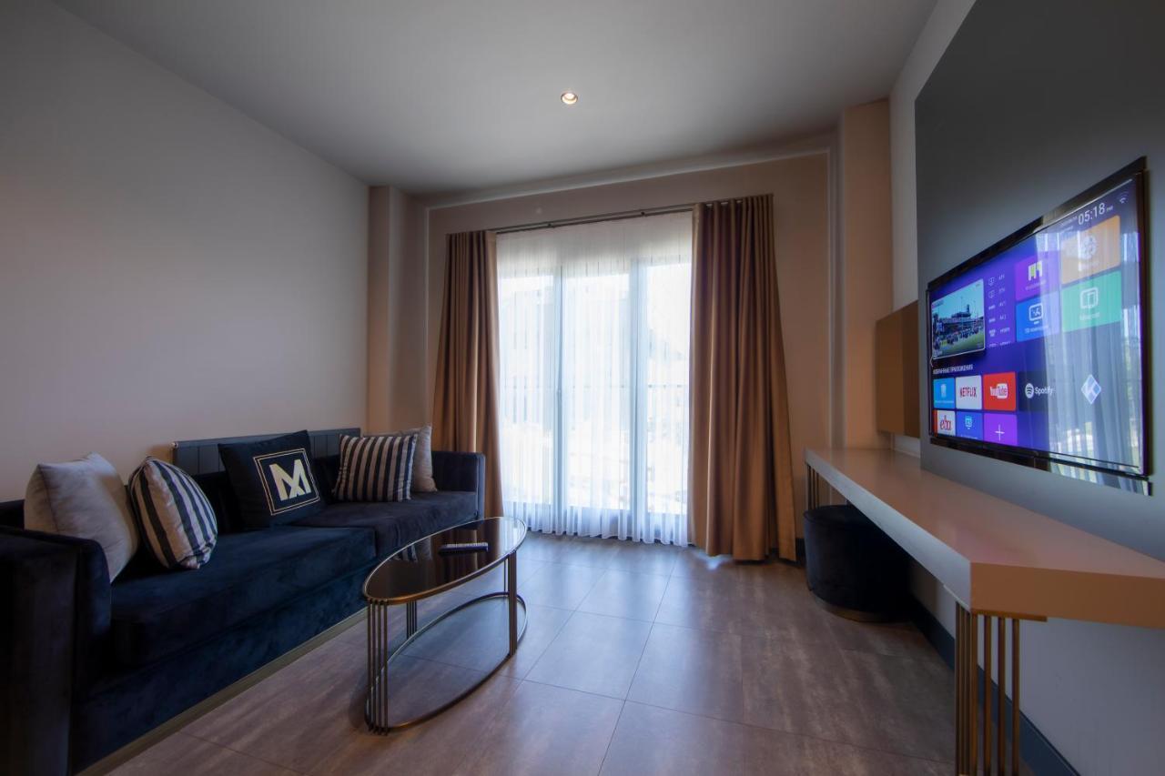 Menalo Hotel Premium Istanbul Airport Exterior photo The photo shows a modern living room with a minimalist design. There's a dark blue sofa adorned with several decorative pillows, one of which has a stylized letter "M." In front of the sofa is a sleek, circular glass coffee table supported by a metal