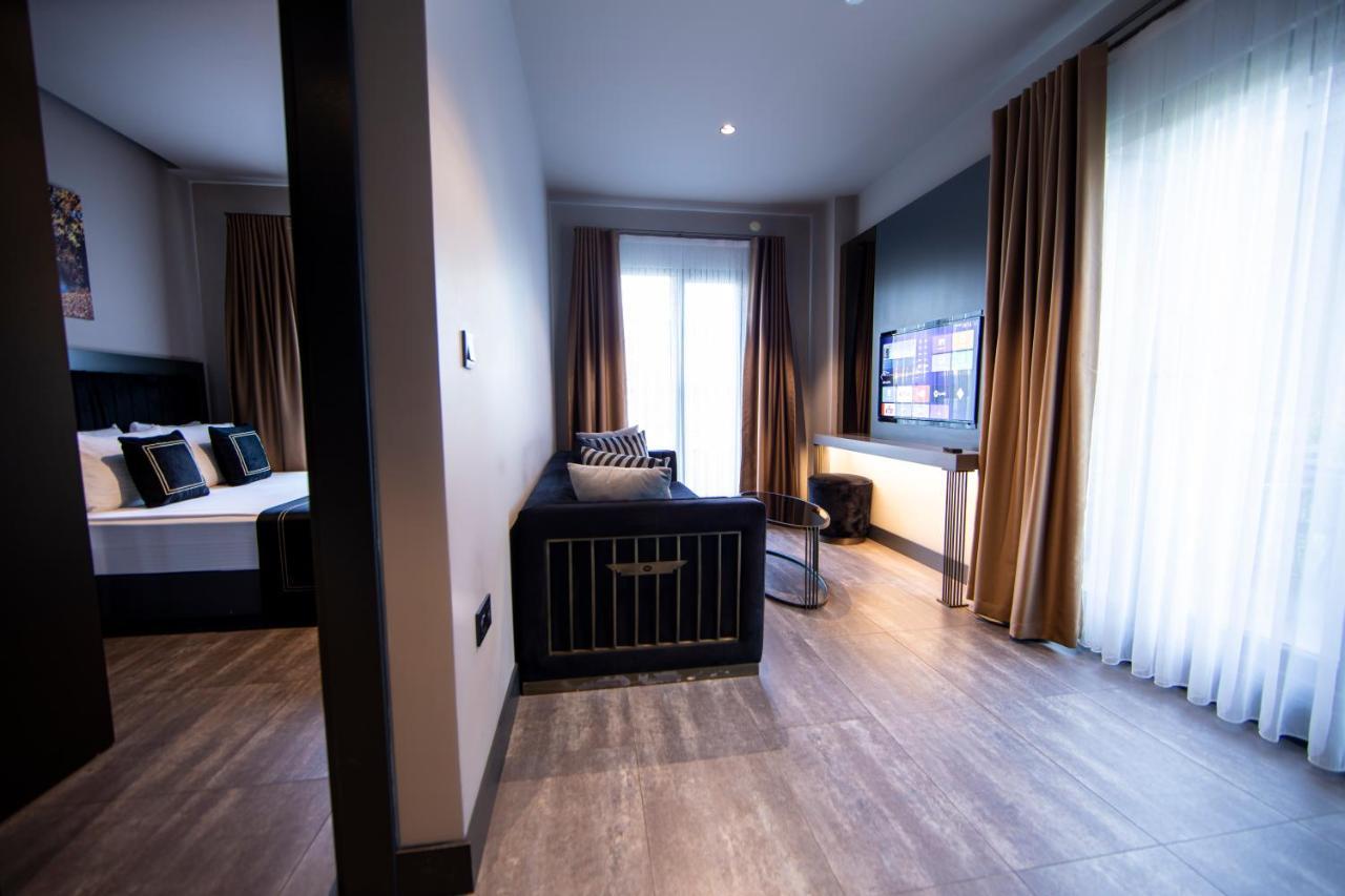 Menalo Hotel Premium Istanbul Airport Exterior photo The photo displays a modern hotel room or suite. On the left side, there is a bed with decorative pillows and curtains, and on the right side, a living area featuring a dark couch or seating arrangement. A television is mounted on the wall in front o