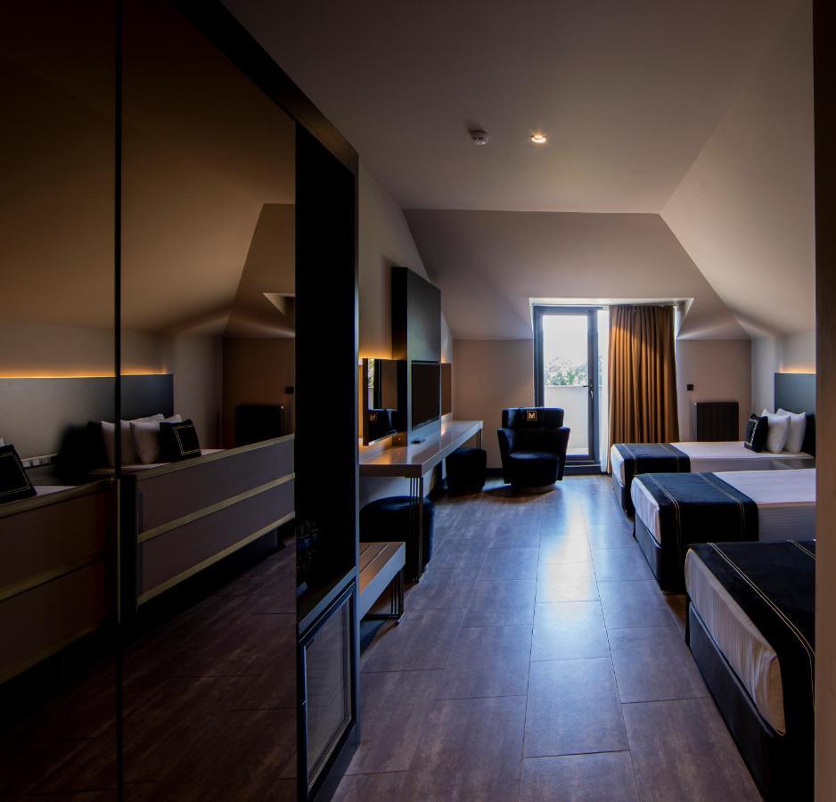 Menalo Hotel Premium Istanbul Airport Exterior photo The photo shows a modern hotel room with a stylish and minimalist design. The room features two beds with dark bedding, a large window with curtains letting in natural light, and a comfortable armchair. There's a sleek desk and furniture, with a mirr