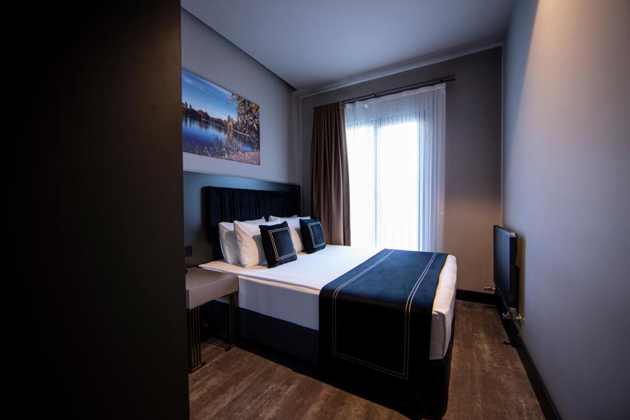 Menalo Hotel Premium Istanbul Airport Exterior photo The photo shows a modern bedroom. It features a double bed with a black headboard and neatly arranged white linens, complemented by dark blue decorative pillows. To the left, there's a piece of artwork depicting a scenic landscape. The large window, 