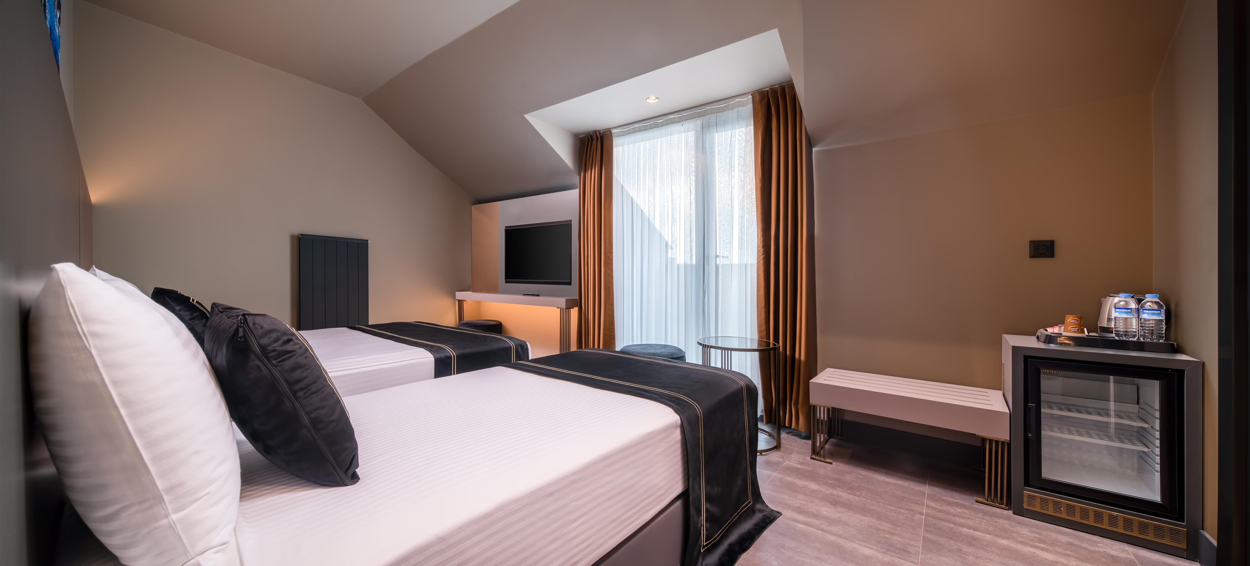 Menalo Hotel Premium Istanbul Airport Exterior photo The photo shows a stylish hotel room featuring two beds with dark-colored throws. There's a large window with sheer curtains allowing natural light to enter, and it is adorned with orange-brown drapes. A flat-screen TV is mounted on the wall opposite